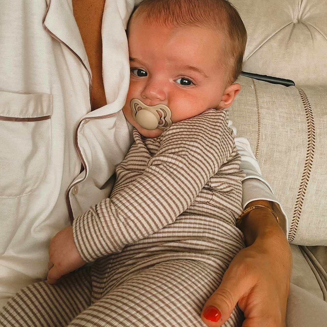 Charles is registered to the contest to win money with this photo: adult_hand, baby, blinds, child, close_up, comfort, couch, face, holding, home, indoor, necklace, pacifier, pajamas, person, relaxation, seated, skin, striped_pajamas, window