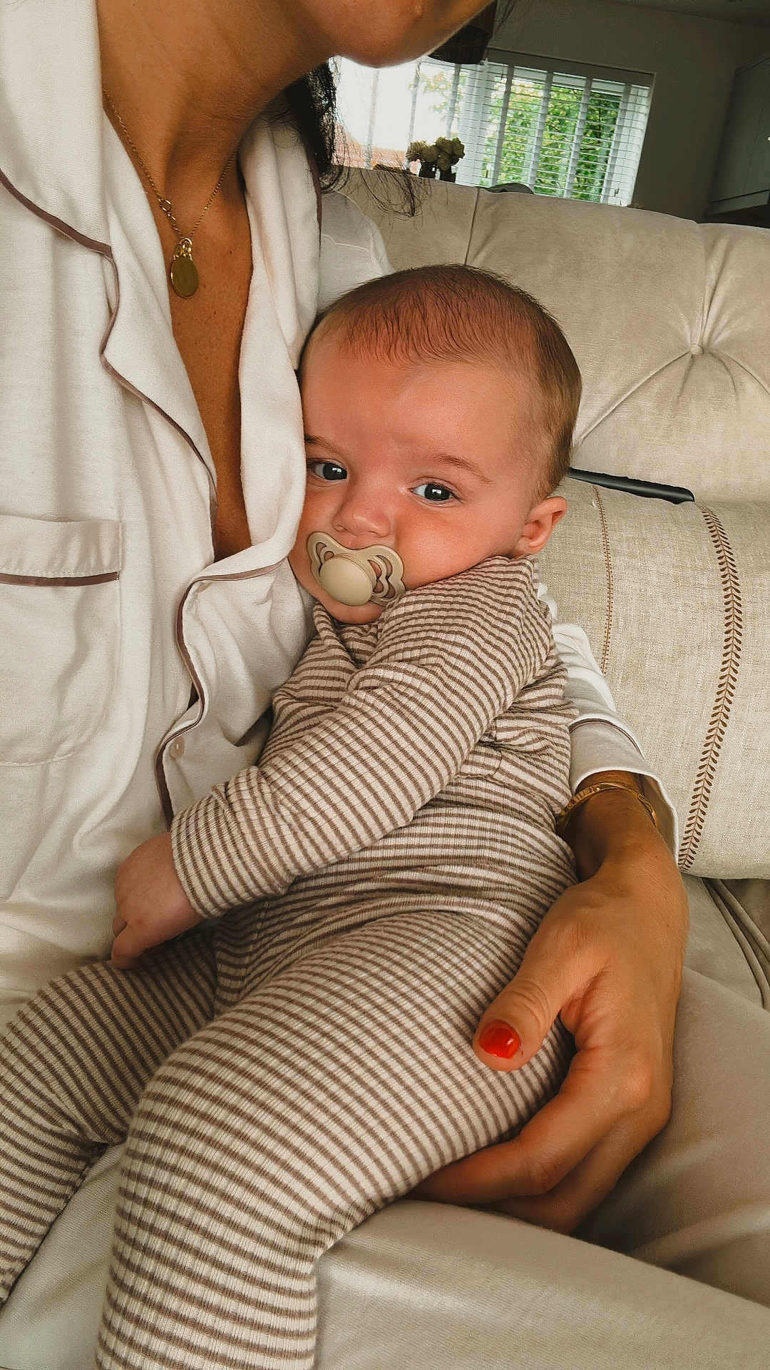 Charles is registered to the contest to win money with this photo: adult_hand, baby, blinds, child, close_up, comfort, couch, face, holding, home, indoor, necklace, pacifier, pajamas, person, relaxation, seated, skin, striped_pajamas, window