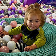 Matilda is registered to the contest to win money with this photo: toddler, child, indoor_play_area, ball_pit, colorful_balls, knitted_nest, turquoise, purple, yellow, playground, curly_hair, striped_pants, green_shirt, smiling, cute, young_child, fun, play, portrait, happy