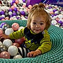 toddler, child, indoor_play_area, ball_pit, colorful_balls, knitted_nest, turquoise, purple, yellow, playground, curly_hair, striped_pants, green_shirt, smiling, cute, young_child, fun, play, portrait, happy