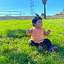 toddler, child, grass, outdoor, sunny, blue_sky, orange_top, pants, shoes, hair_ponytail, tree, palm_tree, power_lines, fence, field, daylight, sitting, nature, person, curious
