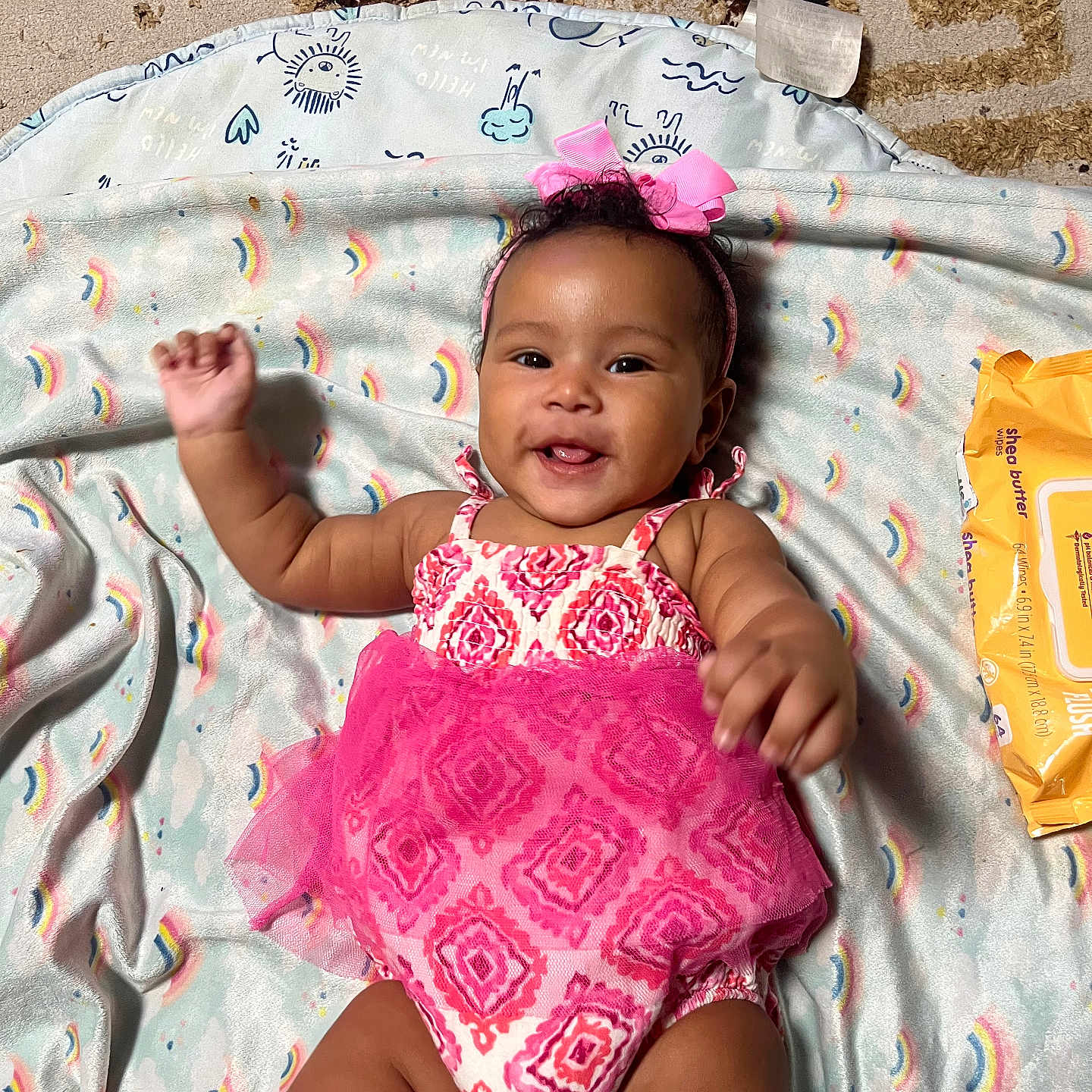 Naziyah is registered to the contest to win money with this photo: baby, blanket, carpet, cheerful, child, cute, happy, headband, indoor, infant, lying_down, person, pink_bow, pink_dress, playful, rainbow_pattern, smiling, toddler, waving_hands, wipes