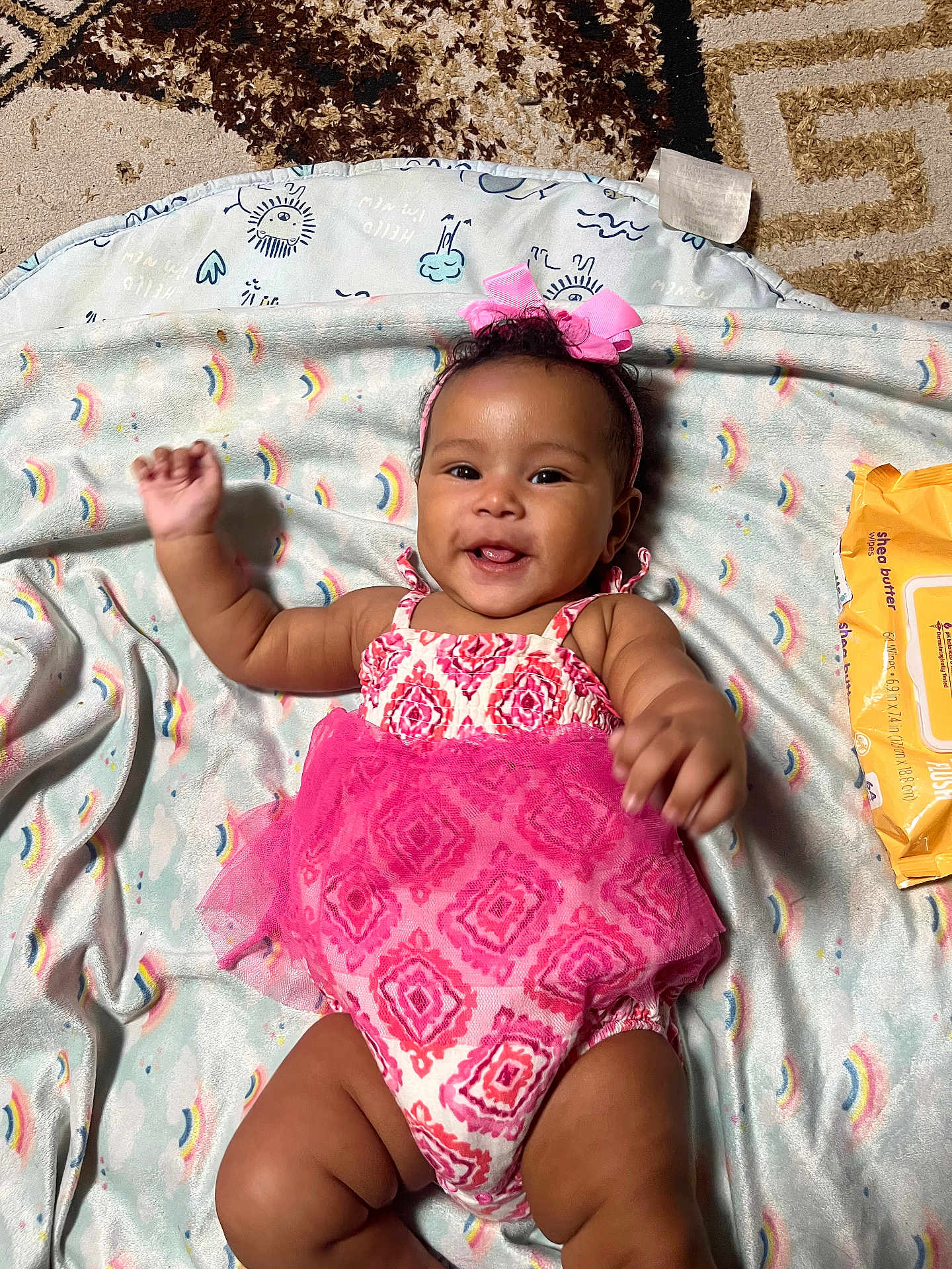 Naziyah is registered to the contest to win money with this photo: baby, child, smiling, pink_bow, pink_dress, blanket, rainbow_pattern, waving_hands, wipes, carpet, indoor, cute, happy, person, infant, headband, toddler, playful, lying_down, cheerful
