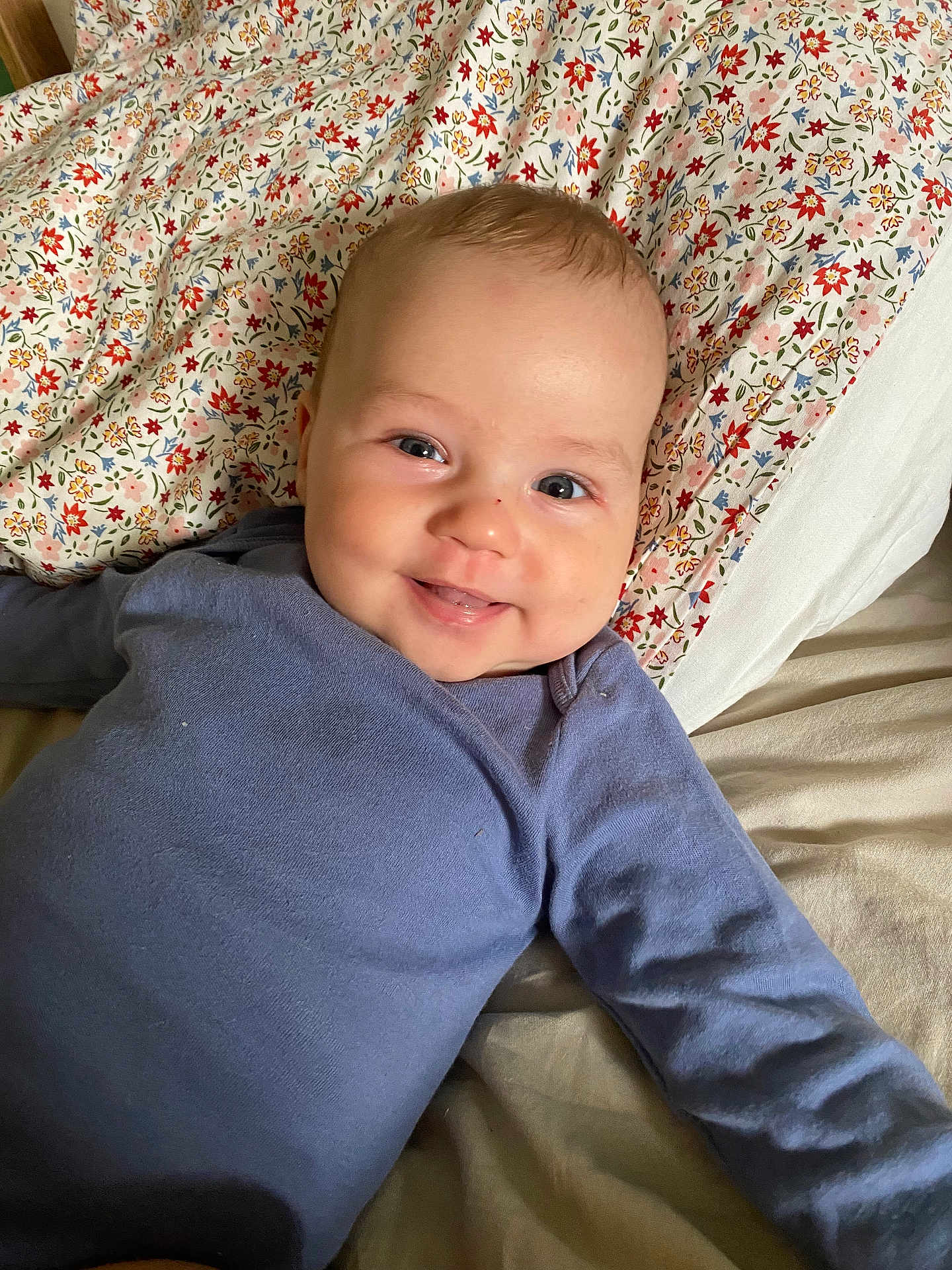 Manoë participe au concours pour gagner de l'argent avec cette photo : baby, smile, blue_clothing, bed, floral_sheet, happy, child, infant, face, skin, person, lying_down, cute, indoors, young_child, head, eyes, cheeks, long_sleeve, comfortable