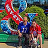 Galen Erso is registered to the contest to win money with this photo: people, dog, jersey, sports_fan, outdoor, sunlight, mascot, sculpture, greenery, storefront, glasses, shorts, sandals, concrete, smiling, posing, white_dog, blue_jersey, red_jersey, reflection