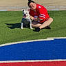 person, dog, artificial_turf, sports_jersey, white_dog, gray_shirt, outdoor, sunny, grass, blue_stripe, red_stripe, pavement, smiling, sitting, pet, tongue_out, casual, happy, playful, daylight