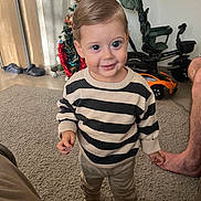 Kasem participe au concours pour gagner de l'argent avec cette photo : toddler, child, striped_sweater, indoor, carpet, christmas_tree, toy_car, footwear, smile, standing, person, pants, sock, hair, furniture, home, decor, floor, leg, adult_foot