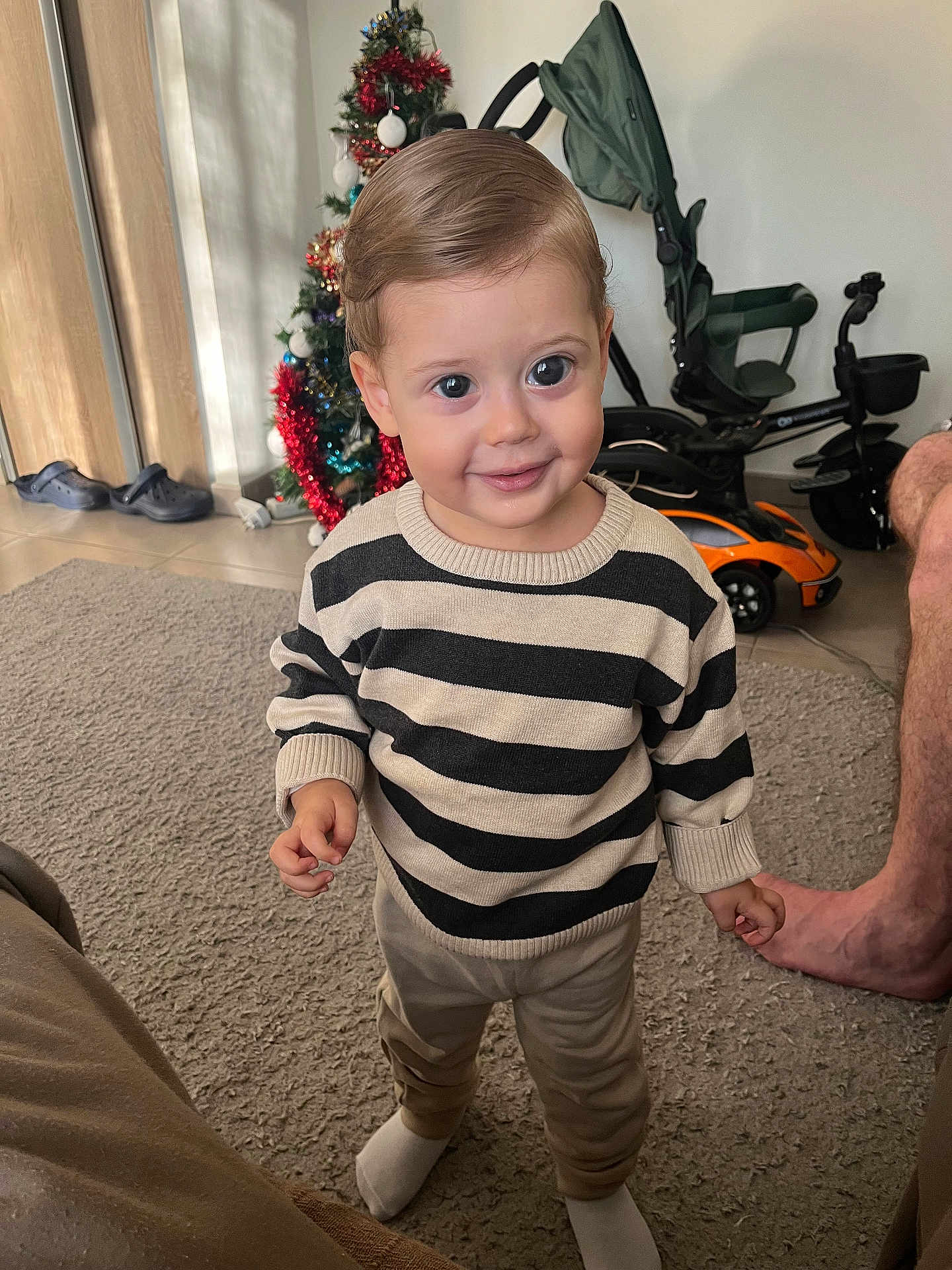 Kasem participe au concours pour gagner de l'argent avec cette photo : toddler, child, striped_sweater, indoor, carpet, christmas_tree, toy_car, footwear, smile, standing, person, pants, sock, hair, furniture, home, decor, floor, leg, adult_foot