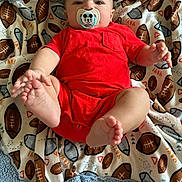Merle joined the competition — help win amazing prizes! baby, infant, pacifier, red_clothing, football_pattern, blanket, feet, hand, lying_down, cute, child, young_child, indoors, soft_texture, smiling_eyes, cozy, comfort, portrait, closeup, playful