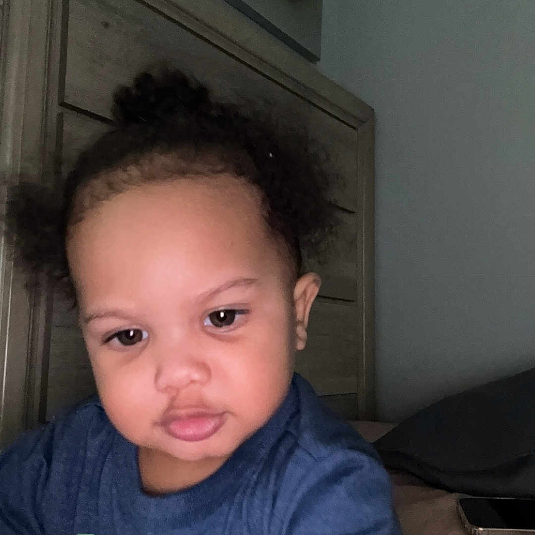 Jaquavion joined the competition — help win amazing prizes! toddler, child, indoor, bedroom, blue_shirt, curly_hair, face, person, headboard, phone, furniture, young_child, serious_expression, skin, arm, closeup, casual_clothing, home, interaction, portrait