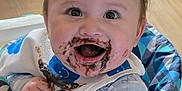 Mavrik is registered to the contest to win money with this photo: baby, child, high_chair, chocolate, messy, happy, smiling, bib, clothing, face, hair, hand, indoors, feeding, cute, infant, sitting, wooden_floor, patterned_clothing, cheerful