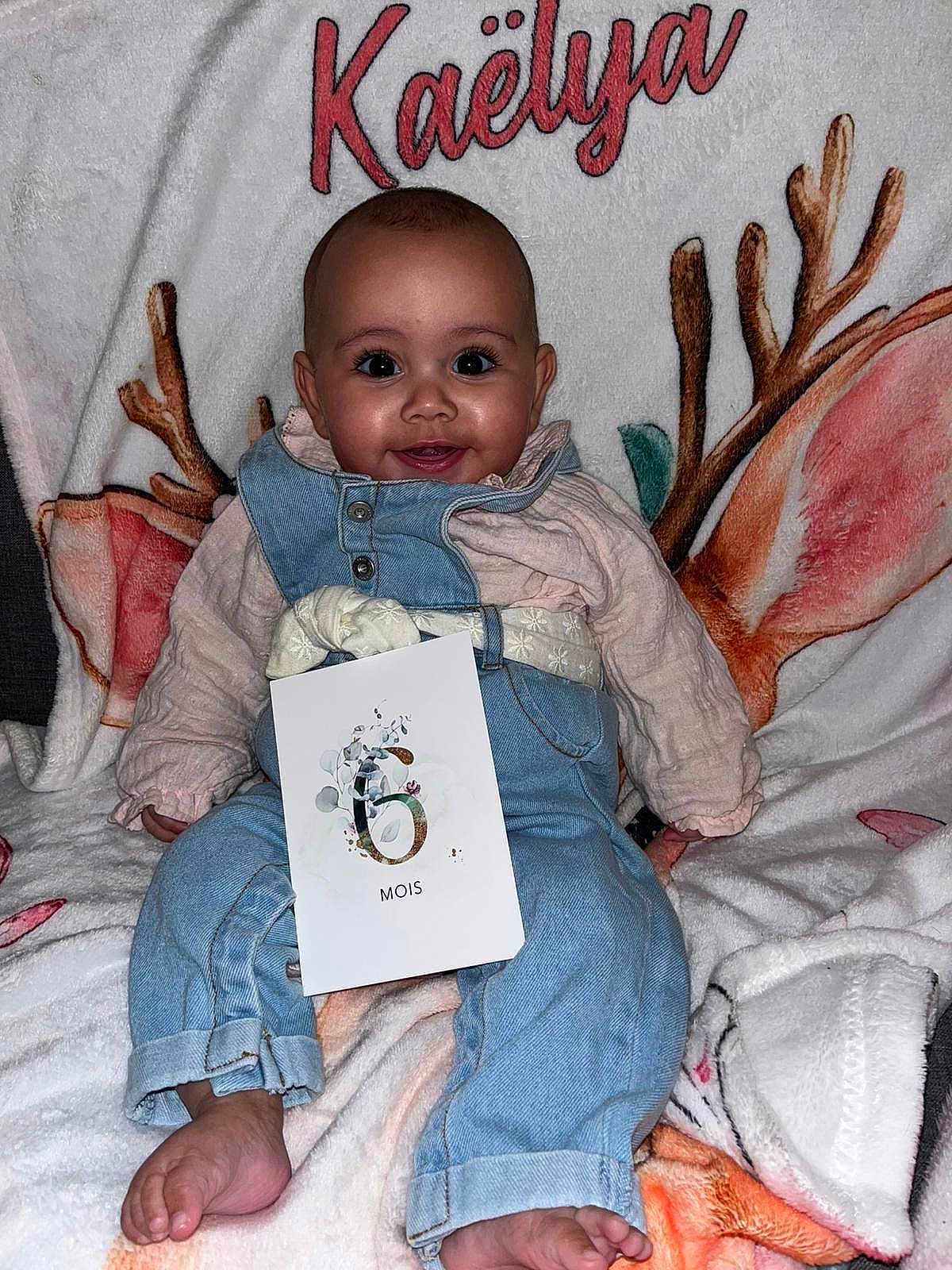 Kaëlya a rejoint le concours — aidez-le/la à gagner de superbes lots ! baby, infant, smile, face, eyes, blanket, name_text, antlers, denim_overall, pink, milestone_card, six_months, card, sitting, feet, portrait, cozy, cute, photography, chubby_cheeks