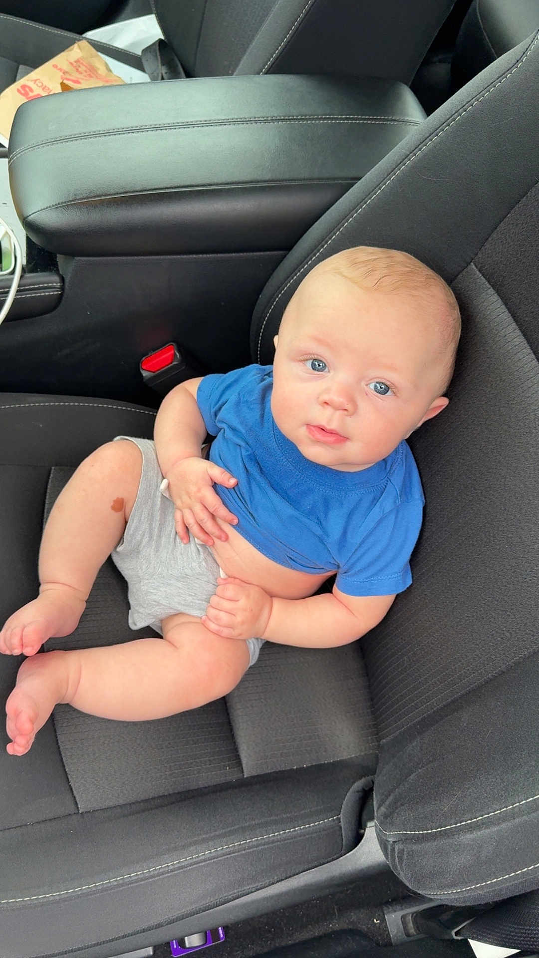 Kaidyn joined the competition — help win amazing prizes! baby, child, car_seat, blue_shirt, gray_shorts, infant, person, cute, skin, legs, feet, hand, seatbelt, car_interior, curious, sitting, young, portrait, casual, relaxed