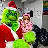 baby, balloons, black_bags, child, christmas, costume, decorations, festive, flooring, green_fur, grinch, holiday, indoor, pacifier, person, pink_bows, playful, red_suit, wall_art, whiteboard