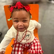 Myamore is registered to the contest to win money with this photo: toddler, child, red_bow, smiling, orange_chair, red_checkered_pants, white_shirt, necklace, indoor, happy, cute, face, person, clothing, sitting, feet, socks, playful, joy, portrait