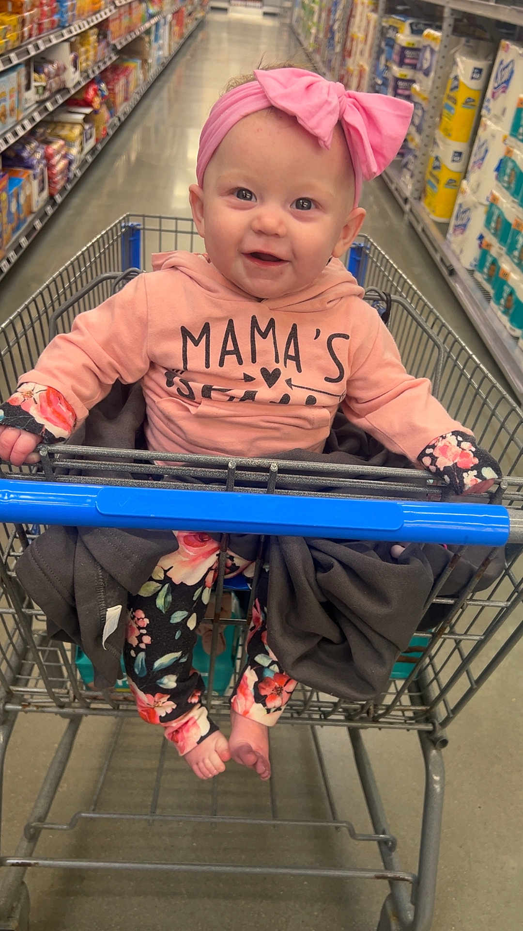 Adaline is registered to the contest to win money with this photo: baby, child, shopping_cart, grocery_store, pink_bow, pink_hoodie, floral_pants, smiling, happy, aisle, store_shelves, barefoot, clothing, infant, headband, person, retail, shopping, cute, indoor