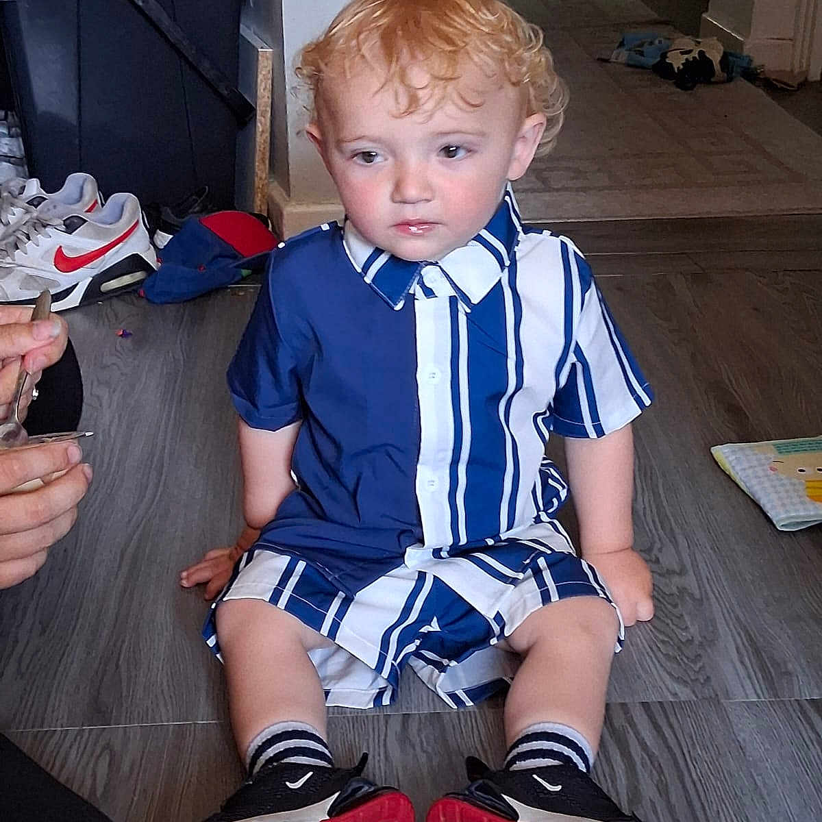 Arlo-Alexander is registered to the contest to win money with this photo: background, black_shoes, blue_and_white, casual_clothing, child, curly_blond_hair, curly_hair, flooring, hand, indoor, person, red_soled_shoes, shoe, sitting, sneakers, socks, striped_clothing, toddler, toy, wooden_floor