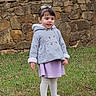child, girl, coat, skirt, tights, grass, stone_wall, outdoor, cat, animal, footwear, white_bow, curly_hair, standing, nature, daylight, casual_clothing, person, young_child, expression