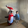 child, girl, indoor, inflatable, penguin, toy, blue_dress, white_tights, shoes, floor, door, window, light, holiday, christmas, decor, smile, standing, wall, home