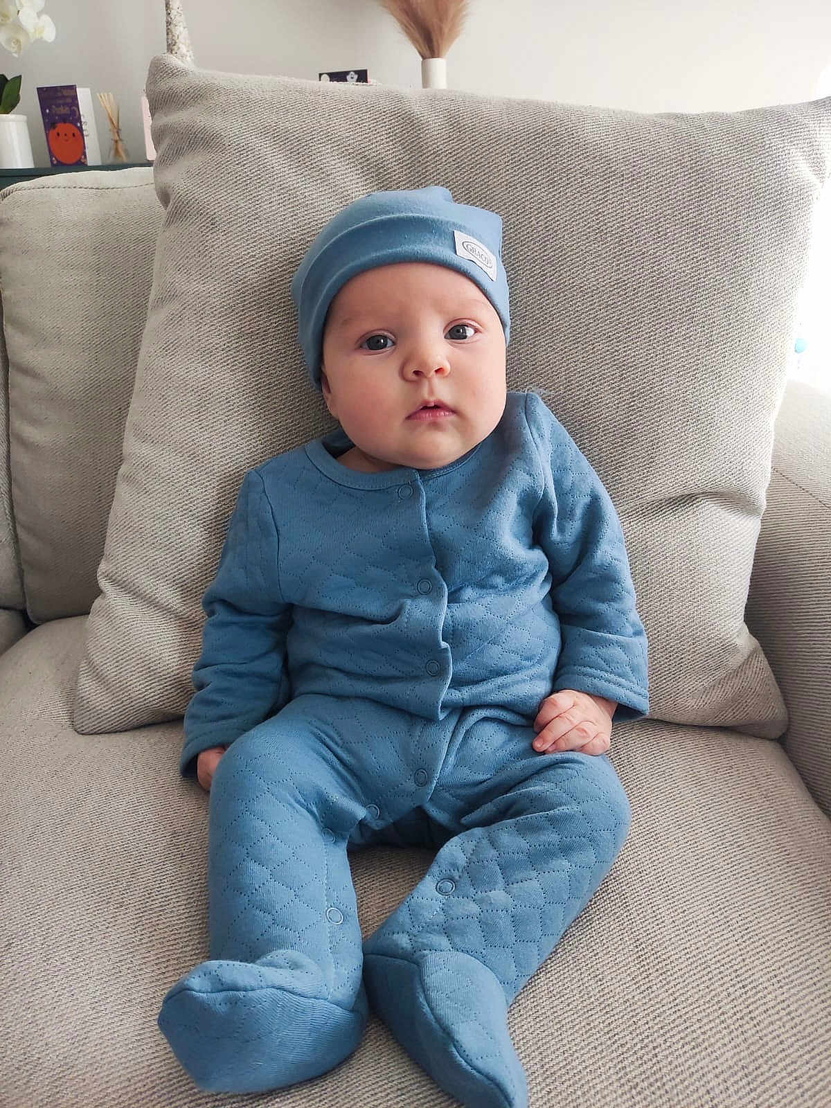 Elias is registered to the contest to win money with this photo: baby, blue_clothing, hat, couch, cushion, infant, indoor, person, portrait, seated, soft_fabric, home_decor, curious_expression, child, face, hands, feet, cozy, quilted, comfortable