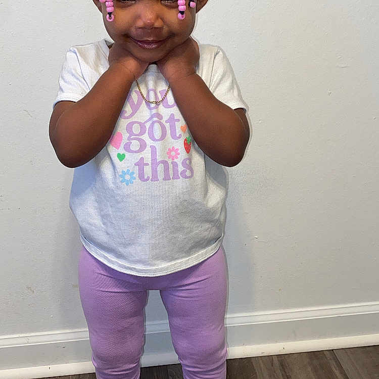 Dalahnni is registered to the contest to win money with this photo: black_sneakers, braided_hair, casual_clothing, child, cute, floor, hands_near_neck, indoors, lavender, leggings, person, pink_beads, portrait, positive_message, smiling, standing, toddler, wall, white_tshirt, wooden_floor