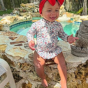 Paisley is registered to the contest to win money with this photo: barefoot, child, fence, greenery, happy, headband, nature, outdoor, playful, pool, portrait, red_bow, smiling, statue, stone_wall, summer, sunlight, swimsuit, toddler, water