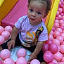 child, toddler, slide, ball_pit, pink_balls, yellow_slide, curly_hair, socks, tshirt, indoor_play_area, playground, cute, seated, looking_at_camera, fun, colorful, plastic_balls, young_child, play, smiling