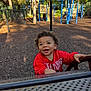 Preston is registered to the contest to win money with this photo: toddler, child, playground, red_sweatshirt, curly_hair, smiling, happy, outdoor, daylight, play_equipment, metal_structure, trees, fence, wood_chips, person, casual_clothing, recreation, fun, young_child, park