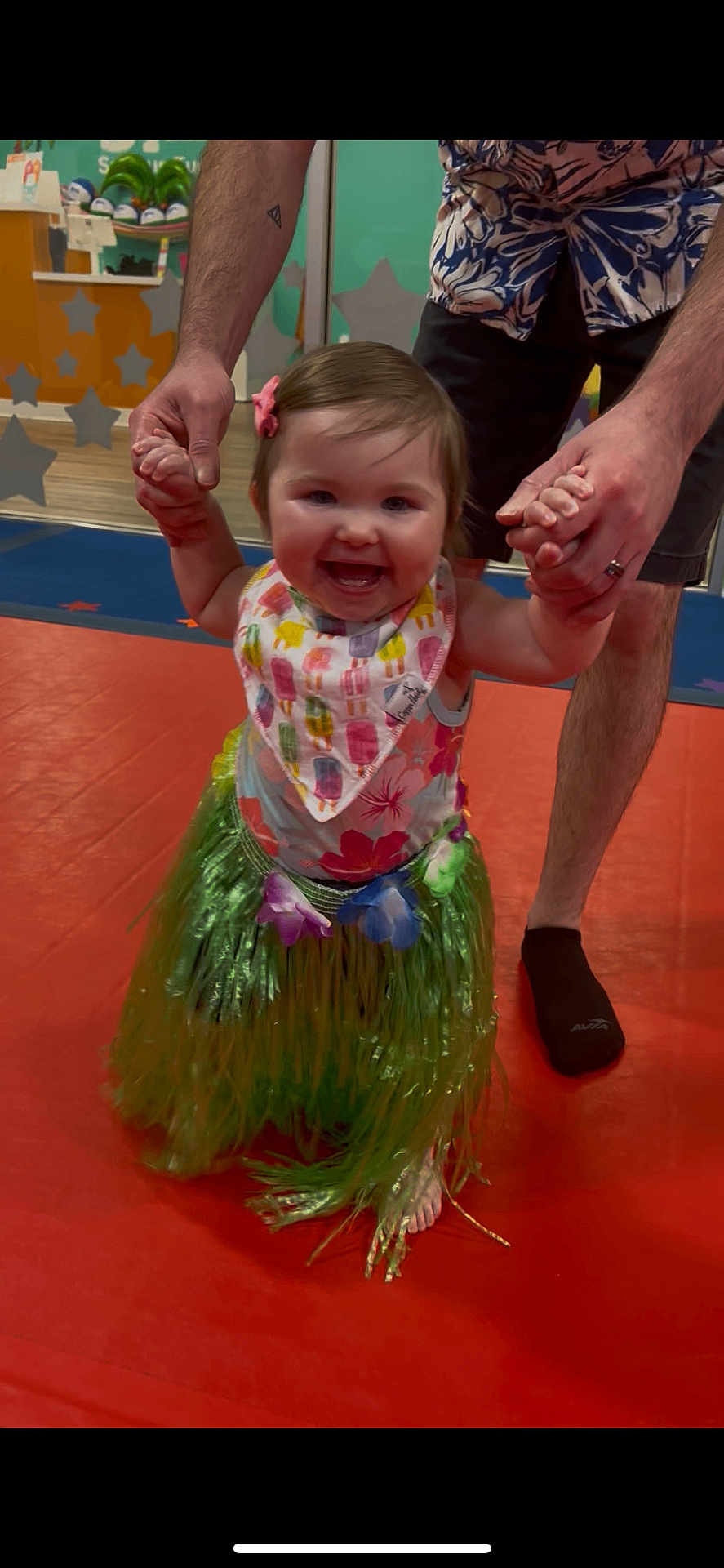 Nora joined the competition — help win amazing prizes! toddler, child, adult, holding_hands, grass_skirt, floral_top, smiling, indoor, play_area, barefoot, colorful, happy, person, floor_mat, tattoo, sock, learning_to_walk, fun, celebration, family