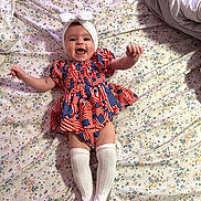 Tatum joined the competition — help win amazing prizes! baby, infant, smiling, happy, dress, headband, bow, socks, lying_down, bed, floral_bedspread, patterned_clothing, white_clothing, cute, child, person, indoors, portrait, cheerful, small_child
