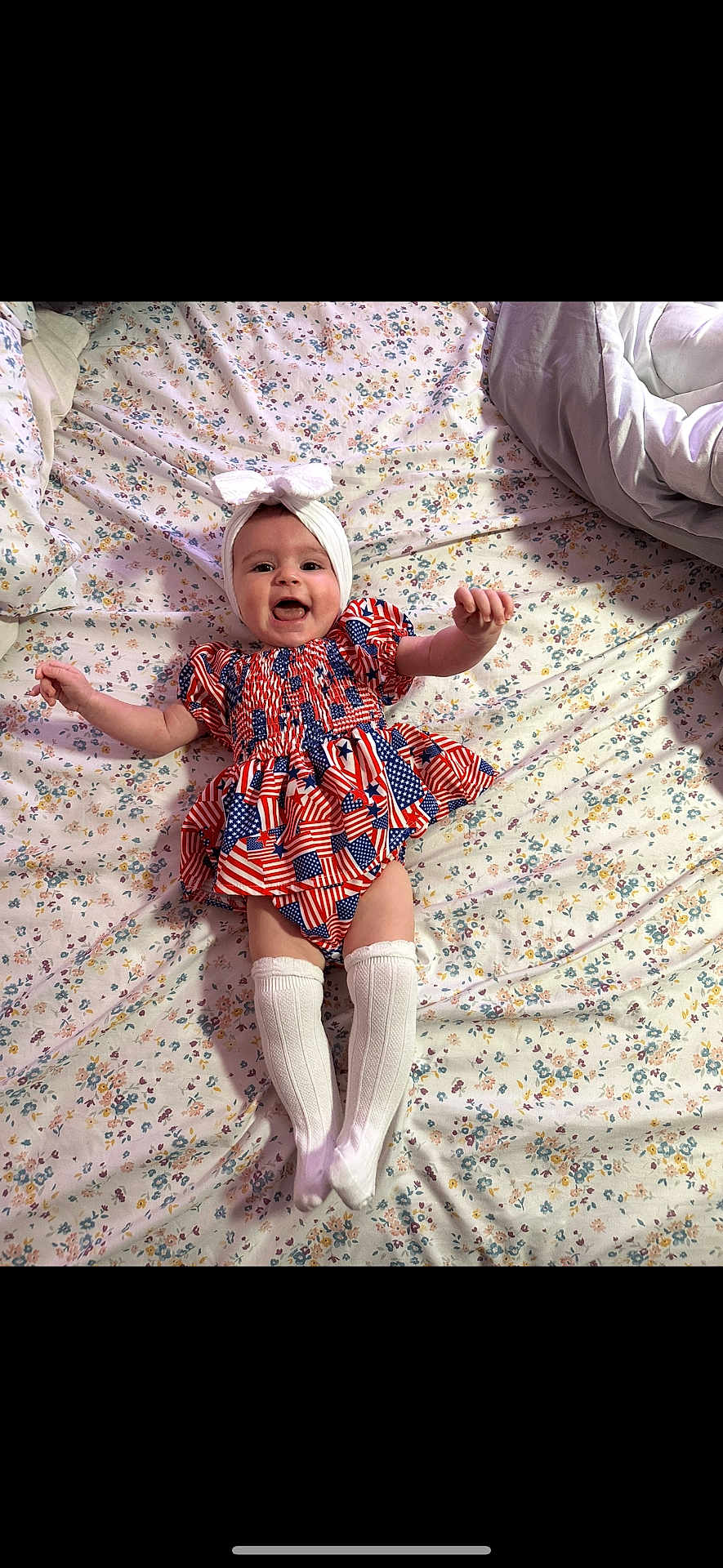 Tatum joined the competition — help win amazing prizes! baby, infant, smiling, happy, dress, headband, bow, socks, lying_down, bed, floral_bedspread, patterned_clothing, white_clothing, cute, child, person, indoors, portrait, cheerful, small_child
