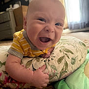 Lucas is registered to the contest to win money with this photo: baby, smile, infant, yellow_clothing, pillow, floral_pattern, indoor, carpet, happy, cute, face, child, young, person, lying_down, closeup, soft_light, home, nursery, playtime