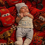 Nathan is registered to the contest to win money with this photo: baby, infant, blanket, plush_toy, car_toy, piston_cup, clothing, hat, socks, face, child, lying_down, red_color, animated_characters, disney_cars, smile, toy, soft_toy, indoor, portrait
