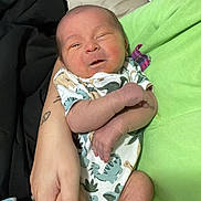 Bowen joined the competition — help win amazing prizes! baby, infant, person, dinosaur_onesie, hand, arm, bed, blanket, clothing, skin, smile, newborn, cute, cozy, human, resting, indoor, sleepwear, fabric, tattoo