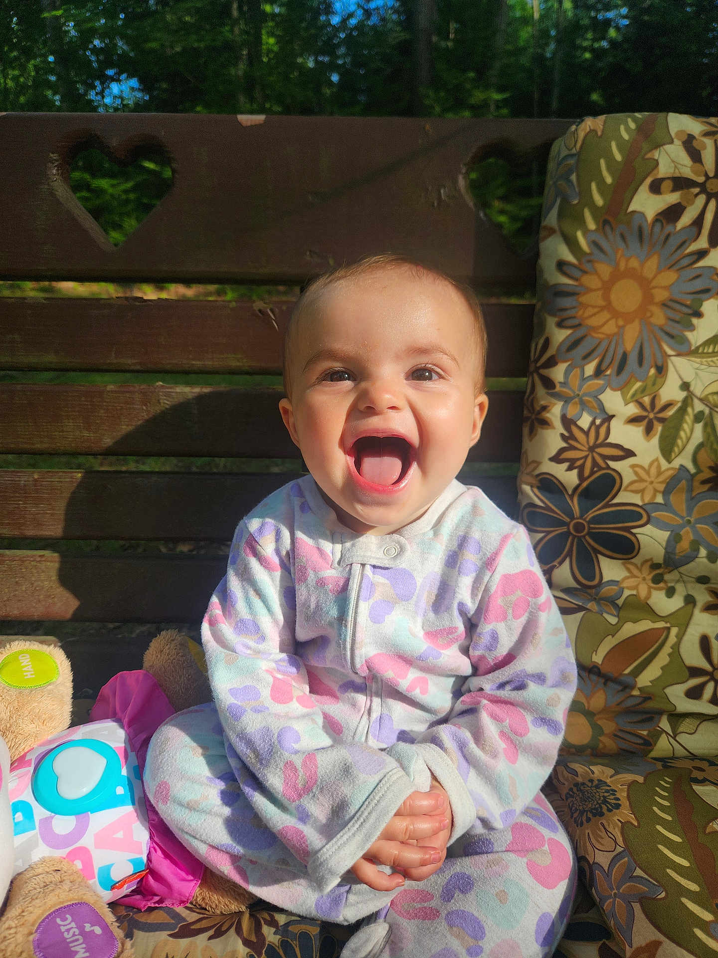 Emma joined the competition — help win amazing prizes! baby, child, smile, laughing, pajamas, bench, wood, heart_shaped_cutout, plush_toy, cushion, floral_pattern, outdoor, sunlight, happy, person, cute, sitting, infant, portrait, colorful