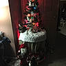cat, christmas_tree, holiday_decorations, santa_figurine, indoor, carpet, table, lights, ornaments, wall, door, decor, festive, animal, pet, curious, dim_lighting, cozy, winter, celebration