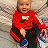 Kasey is registered to the contest to win money with this photo: baby, bed, bedding, black_pants, blanket, child, close_up, cozy, face, happy, indoors, infant, nike, pillow, portrait, red_shirt, sitting, smiling, sock, sportswear