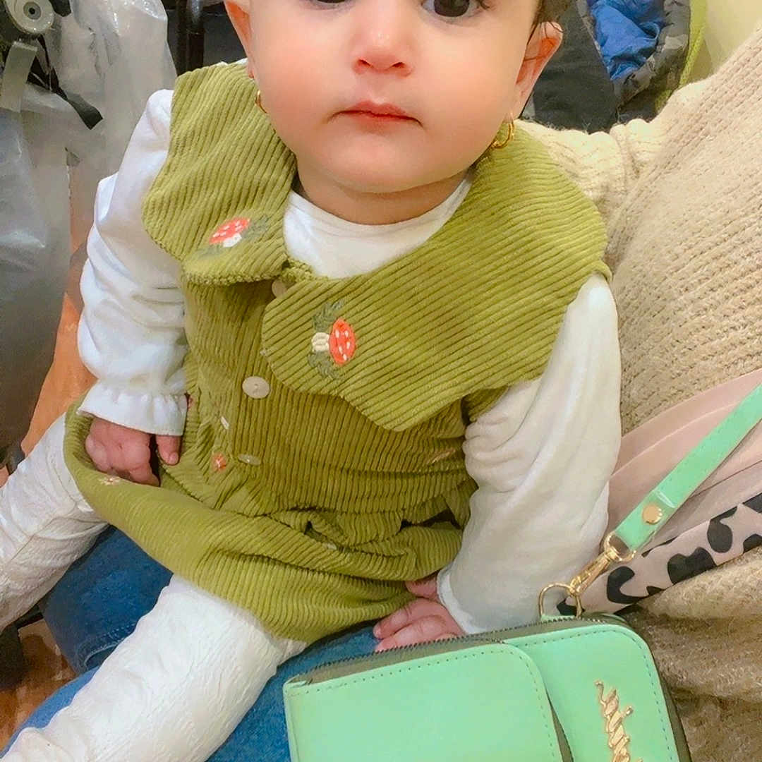 Renad joined the competition — help win amazing prizes! calm, child, curly_hair, cute, earrings, eyes, face, floor, green_dress, handbag, indoor, jeans, long_sleeve, person, purse, sitting, stroller, toddler, white_clothing, wood_floor