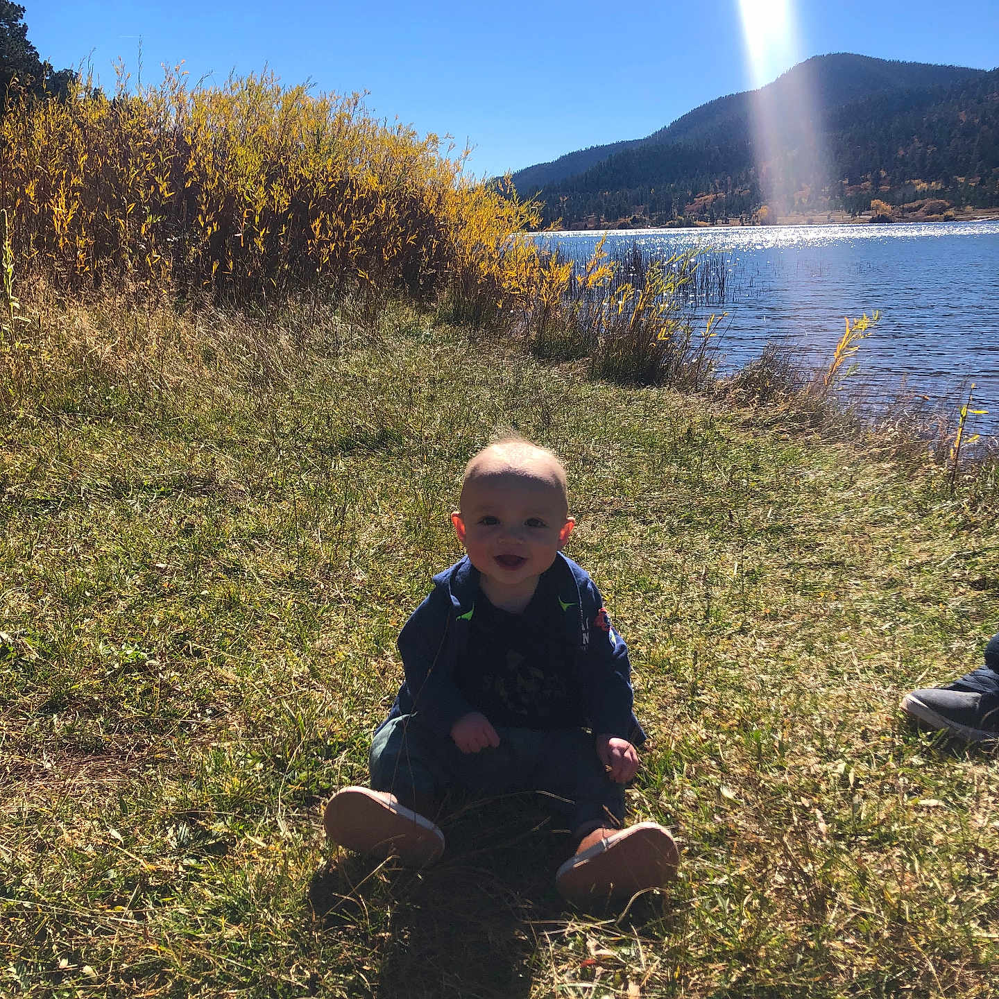 Mason is registered to the contest to win money with this photo: baby, clothing, face, grass, head, lake, nature, outdoors, pants, person, photography, plant, portrait, scenery, sitting, sky, soil, sunlight, vegetation, water
