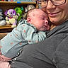 newborn, baby, infant, parent, adult, sleeping, cuddling, portrait, smiling, glasses, nose_ring, onesie, clothing, stuffed_animals, toys, shelf, nursery, indoor, family, closeup
