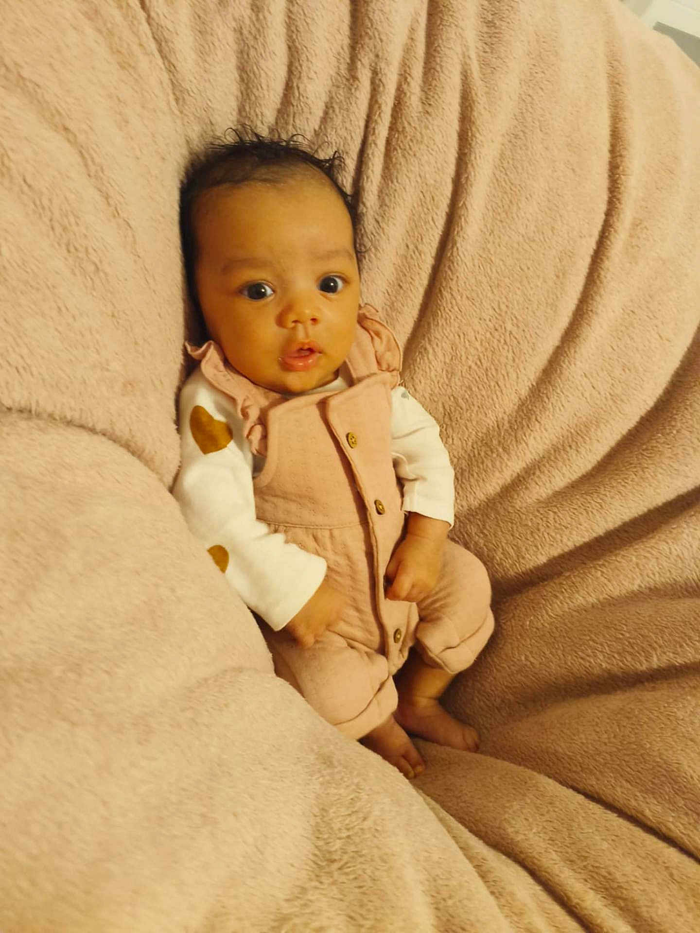 Lya participe au concours pour gagner de l'argent avec cette photo : baby, infant, newborn, pink_overalls, onesie, blanket, soft_texture, cozy, portrait, wide_eyes, curly_hair, skin, hands, toes, seated, bedding, plush, looking_at_camera, cute, indoor