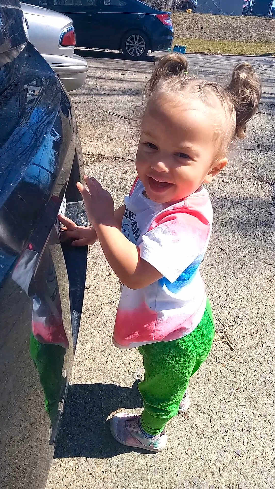 River joined the competition — help win amazing prizes! child, toddler, smiling, standing, outdoor, car, pavement, sunlight, hair_buns, colorful_clothing, shoes, green_pants, white_tshirt, reflection, happy, person, daylight, playful, young_child, cute