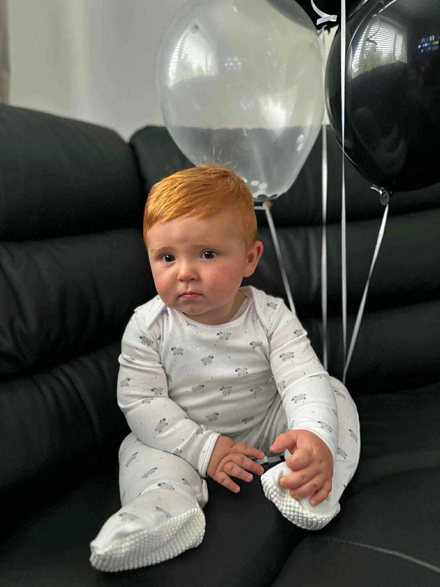 Henry joined the competition — help win amazing prizes! baby, child, red_hair, pajamas, sheep_pattern, black_couch, balloons, white_balloon, black_balloon, indoor, sitting, footwear, cozy, cute, infant, portrait, one_person, expression, hand, looking