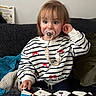 blanket, blue_eyes, book, casual_clothing, child, couch, cozy, curly_hair, home, indoor, pacifier, person, playful, reading, red_bow, sitting, striped_sweater, toddler, toy, young_child