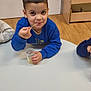 blue_sweatshirt, boy, casual, child, clothing, cup, daylight, face, furniture, holding, indoor, person, plastic_cup, playful, sitting, smile, stick, table, wooden_floor, young