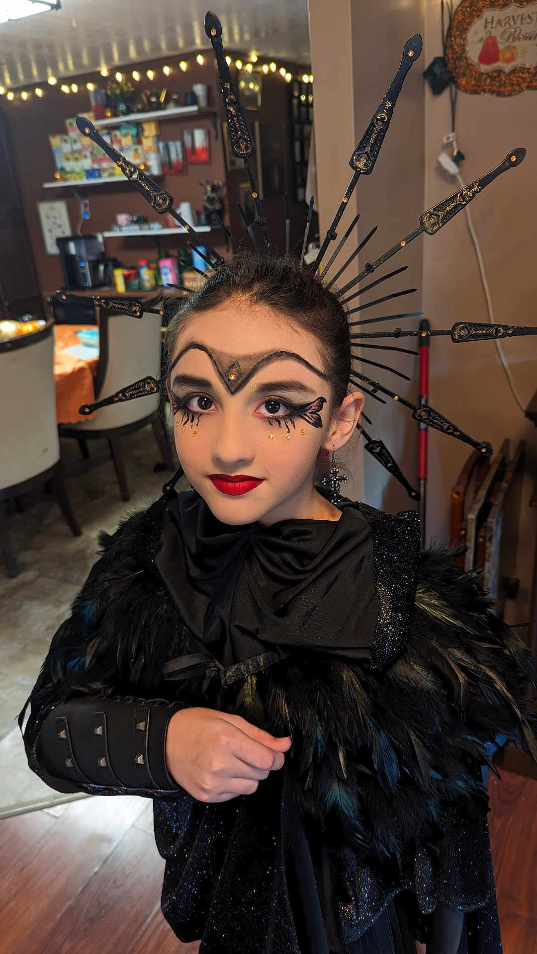 Lilianna is registered to the contest to win money with this photo: child, face_paint, makeup, costume, black_feathers, headpiece, indoor, portrait, young_girl, red_lipstick, decorative_accessory, dramatic_eyeliner, black_clothing, arm_cuff, earring, hand, expression, home_interior, lighting, person