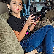 Lilianna is registered to the contest to win money with this photo: child, girl, braids, smartphone, couch, living_room, curious, jeans, black_tshirt, earrings, barefoot, pillow, bookshelf, painting, curtains, home, indoor, casual, relaxed, seated