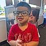 child, boy, glasses, red_shirt, indoor, window, poster, clapping, smiling, short_hair, carpet, advertisement, casual_clothing, daylight, face, person, happy, young_child, hand_gesture, eyewear