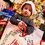 child, toddler, santa_hat, holiday, christmas_tree, gift_bag, presents, festive, holiday_decor, smiling, indoors, celebration, winter, holiday_lights, red, green, white, face, clothing, happy