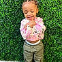 Remi is registered to the contest to win money with this photo: toddler, child, smiling, braided_hair, green_bow, pink_top, olive_pants, white_shoes, leafy_wall, green_background, standing, happy, playful, casual_clothing, young_child, outdoor_style, cute, portrait, fashion, joyful